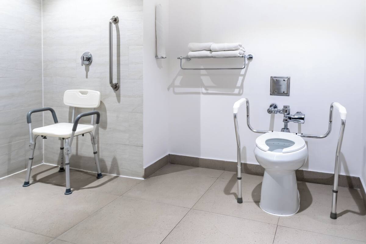 A bathroom with a white toilet, a white sink, and a white shower. There are grab bars next to the toilet and a white chair.