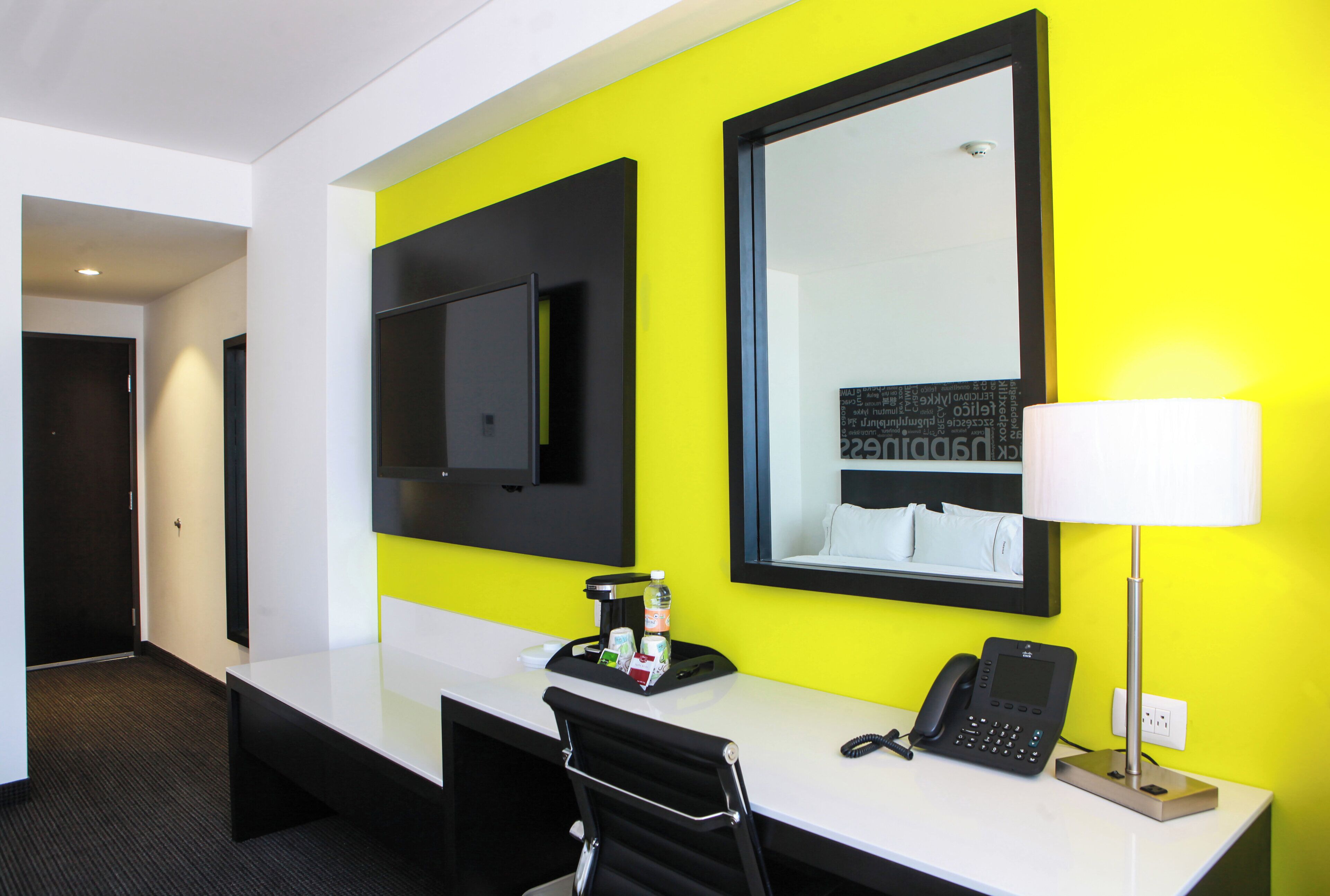 Een hotelkamer met een wit bureau, een zwarte stoel, een flatscreen televisie aan de muur, een bedlampje en een telefoon.