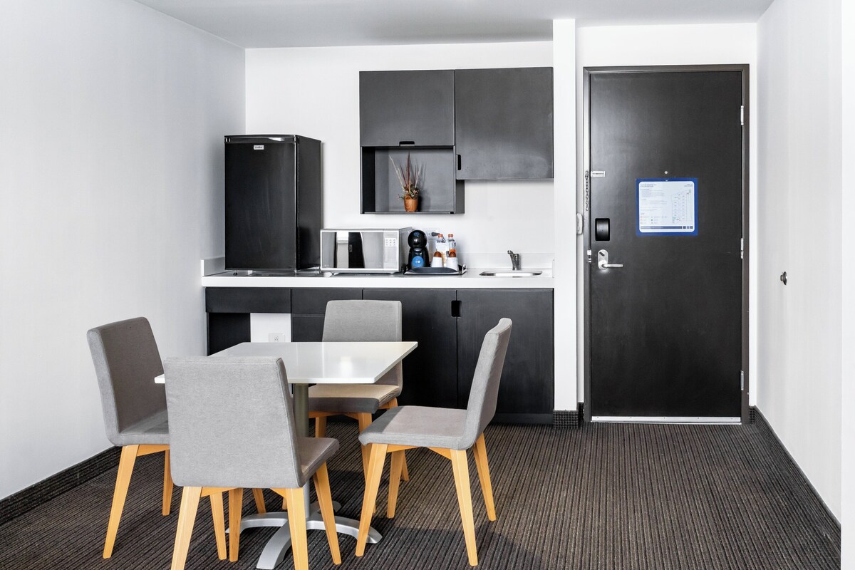 A compact kitchen with a refrigerator, microwave, and coffee maker, a small dining table with four chairs, and a door with a notice.