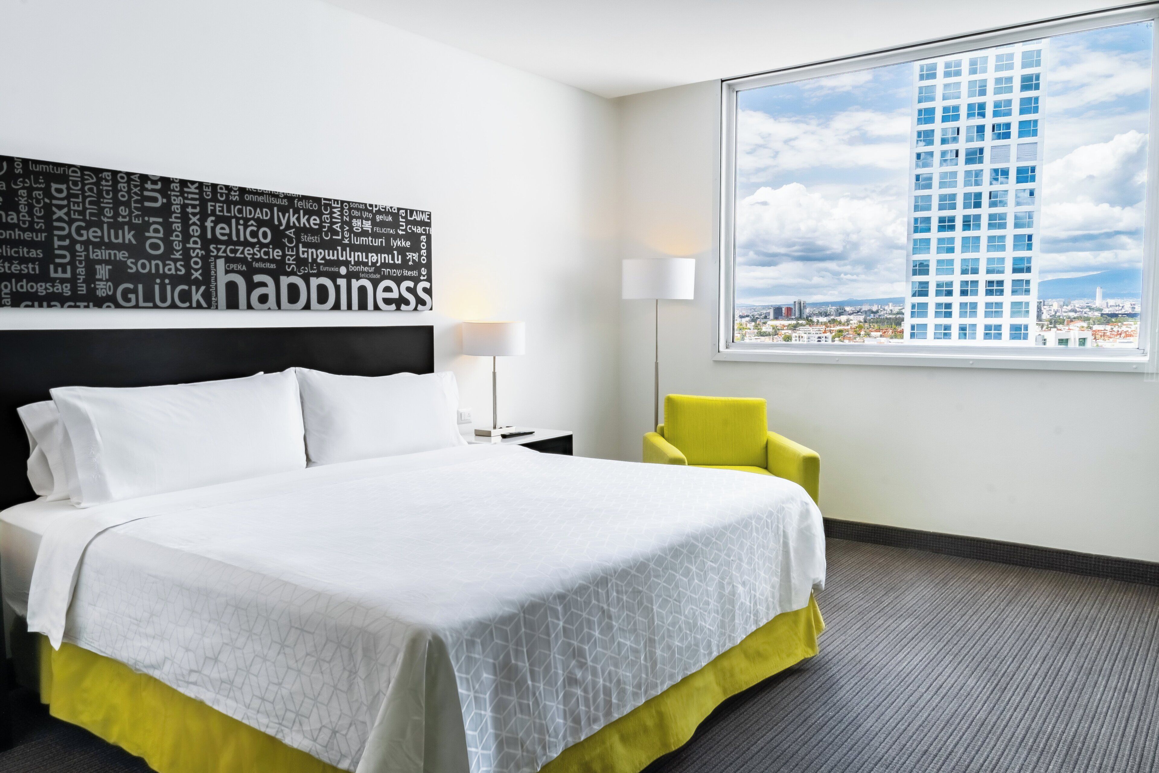 Une chambre d’hôtel avec un lit, un fauteuil jaune, une grande fenêtre donnant sur la ville et un nuage de mots sur le mur.