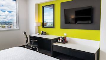 A modern hotel room with a large bed, a desk with a chair, a TV mounted on the wall, and a window offering a city view.