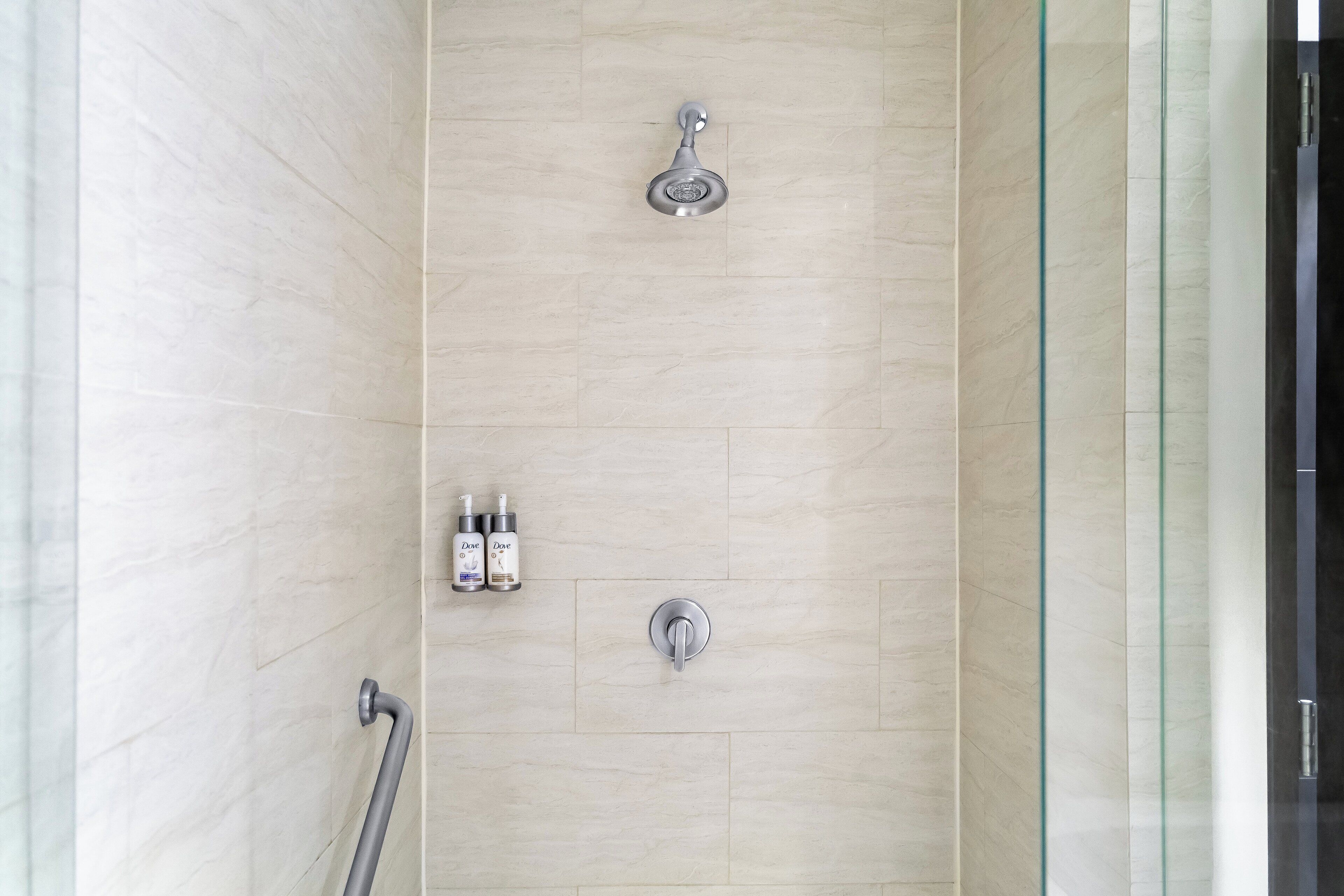 A shower with a handheld showerhead and a wall-mounted faucet, two Dove body wash bottles, and a glass shower enclosure.