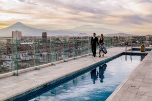 Una piscina en la azotea con barandillas de cristal, vista a una ciudad y una montaña al fondo.