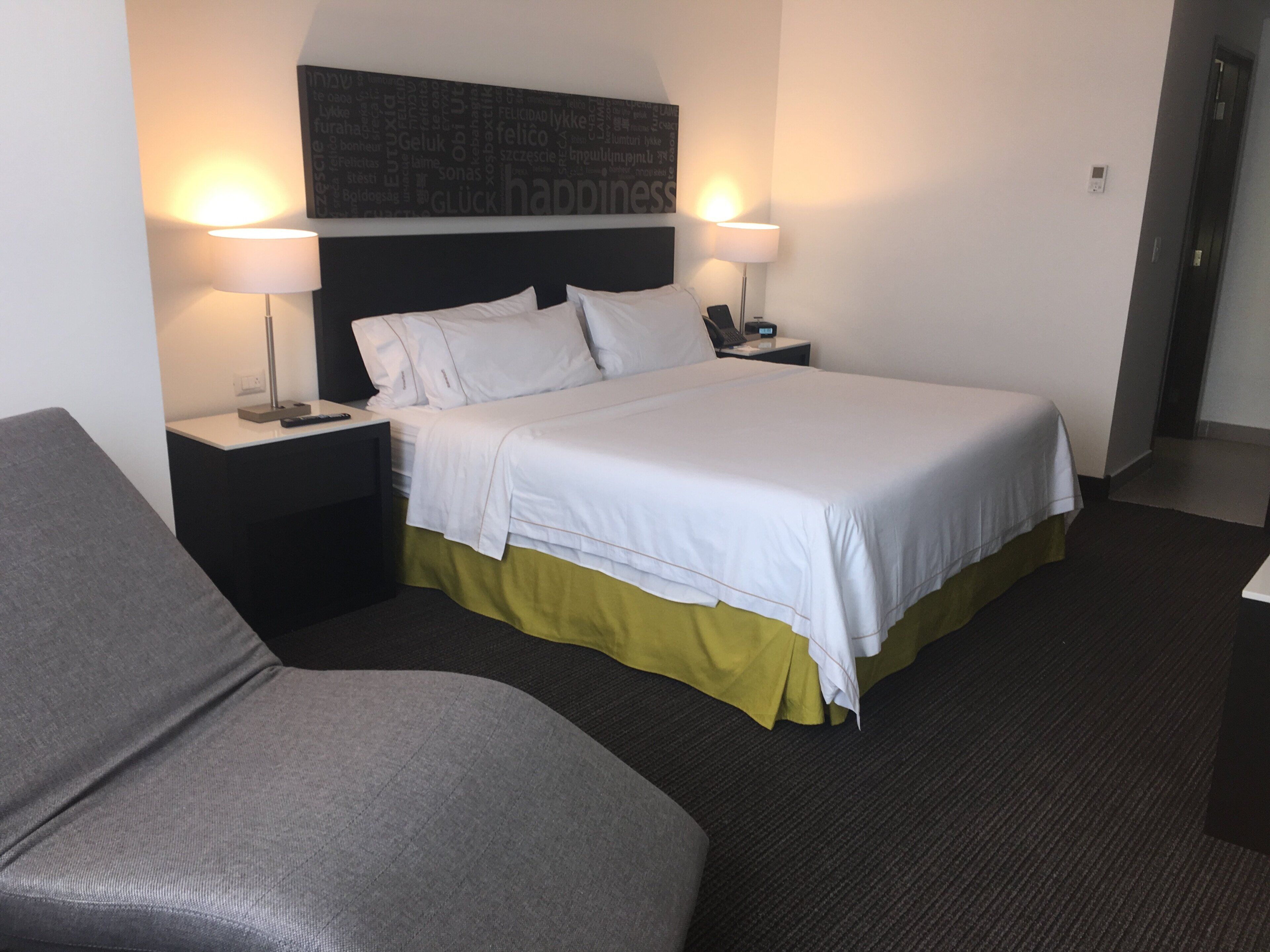 A hotel room with a bed, two bedside lamps, a nightstand, a grey armchair, and a wall with a word cloud.
