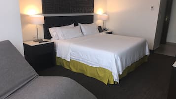 A hotel room with a bed, two bedside lamps, a nightstand, a grey armchair, and a wall with a word cloud.