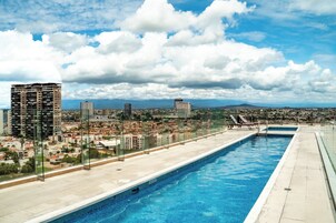 Una piscina en la azotea con vista a la ciudad, barandillas de vidrio y sillas de descanso.