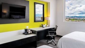 Habitación de hotel con cama, escritorio, silla, televisor, espejo, lámpara y ventana con vista a la ciudad.
