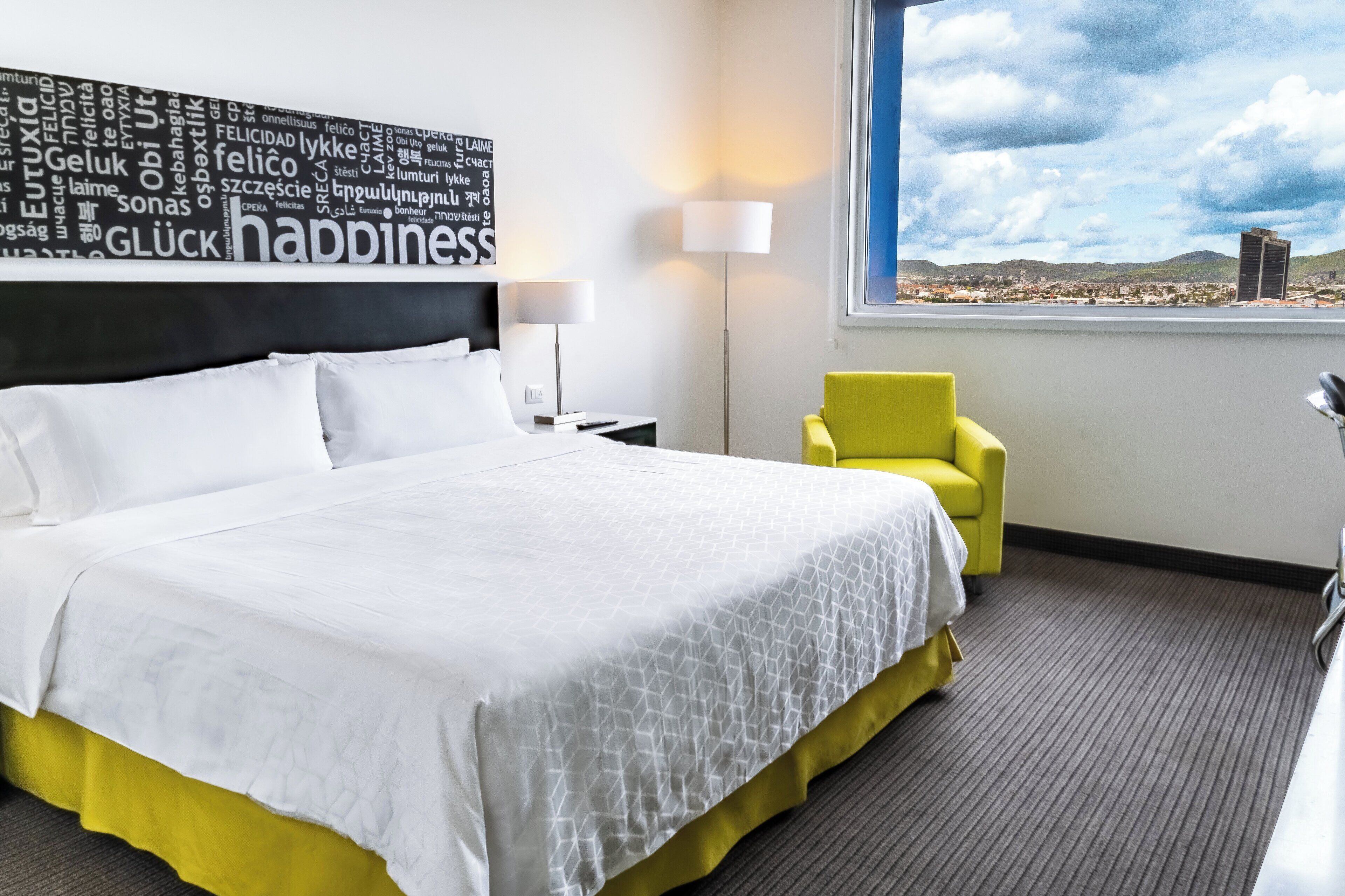 Une chambre d’hôtel avec un grand lit, un fauteuil jaune, une vue sur le paysage urbain et un nuage de mots sur le mur.