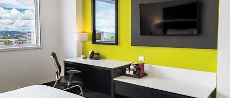 Quarto de hotel moderno com uma cama grande, uma mesa com cadeira, TV na parede e janela com vista para a cidade.