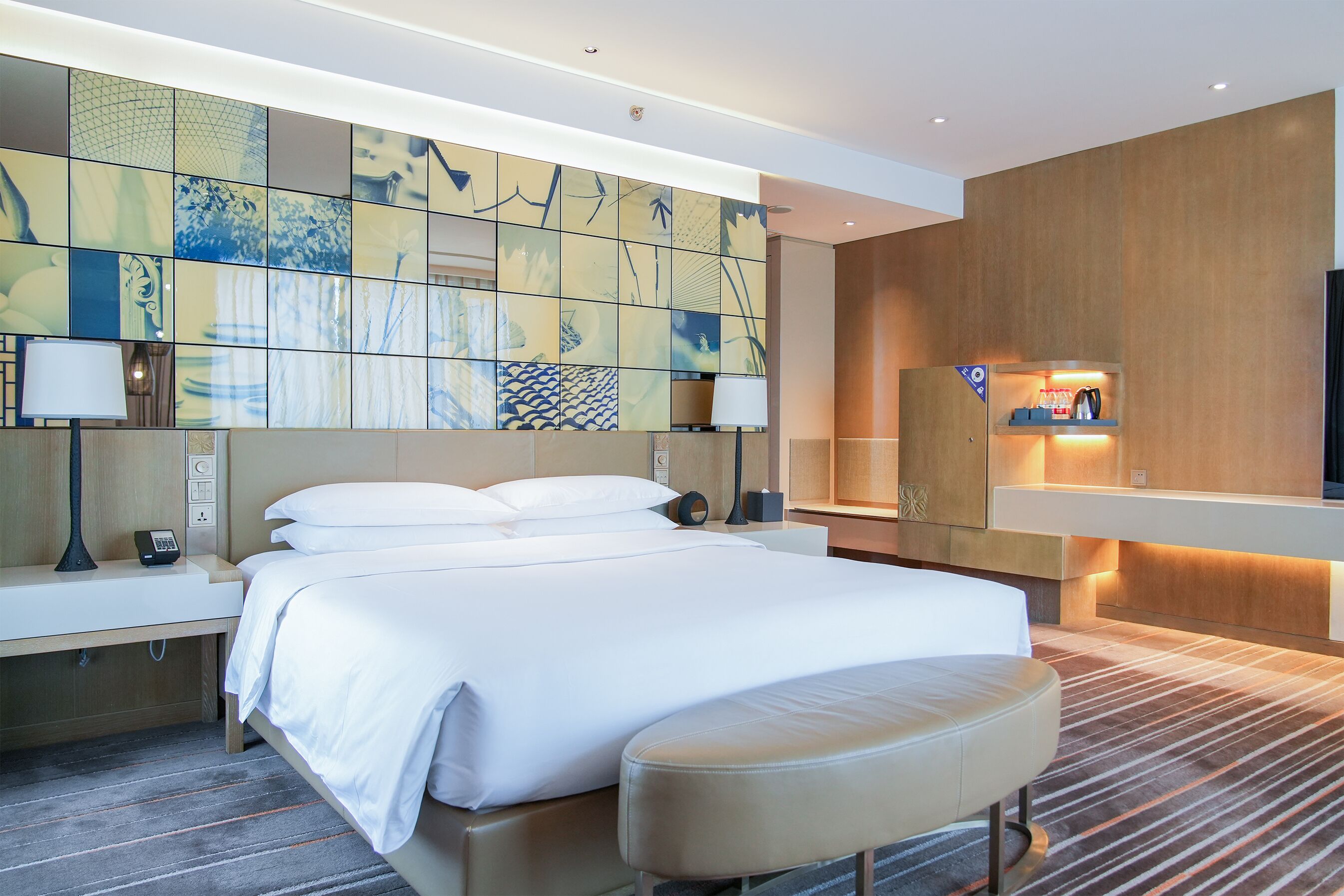 Ett modernt hotellrum med en stor säng, sängbord, ett skrivbord, en lampa, en spegel och en vägg med ett geometriskt kakelmönster.