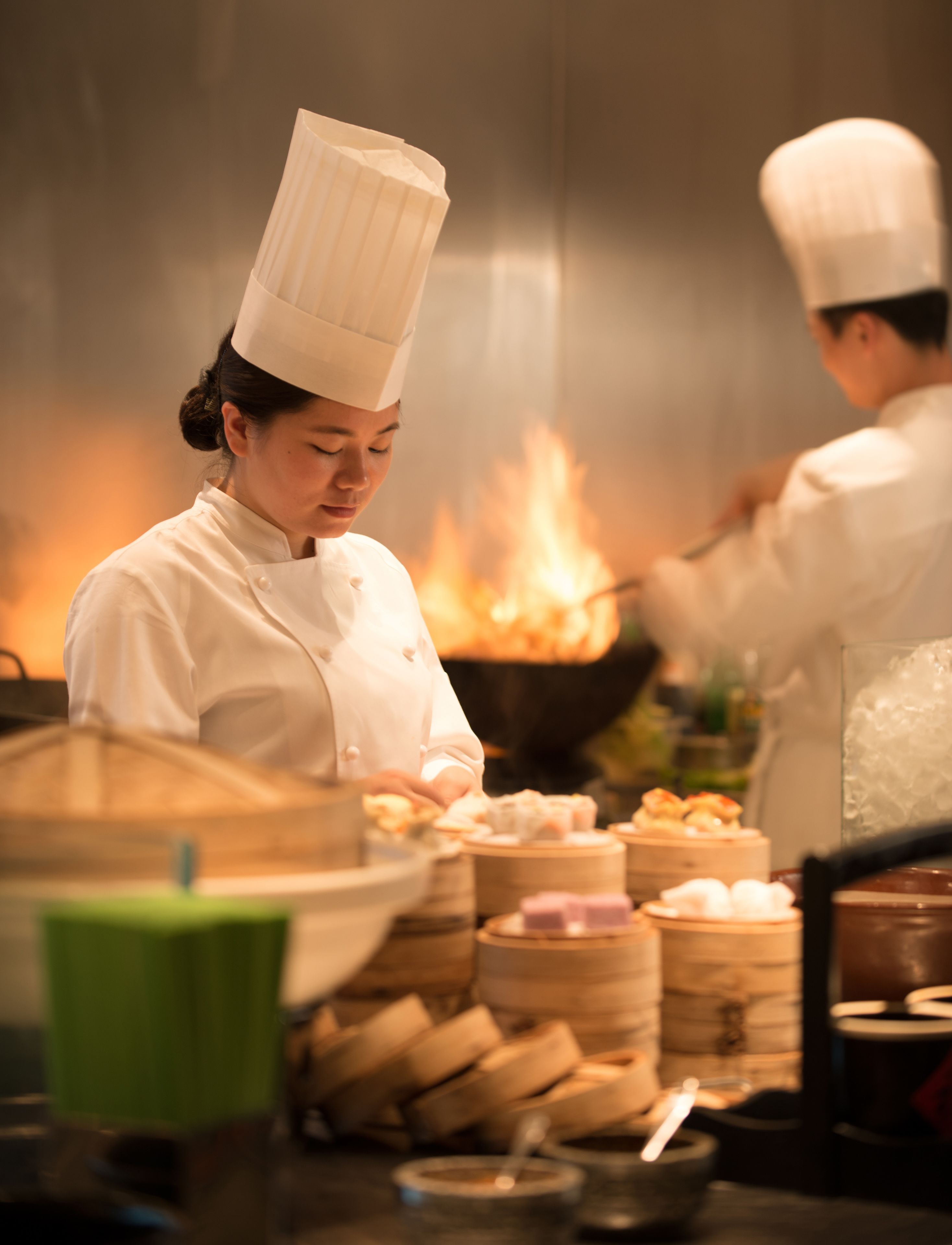 A chef in a white uniform with a tall hat, cooking in a kitchen with a visible fire in the background.