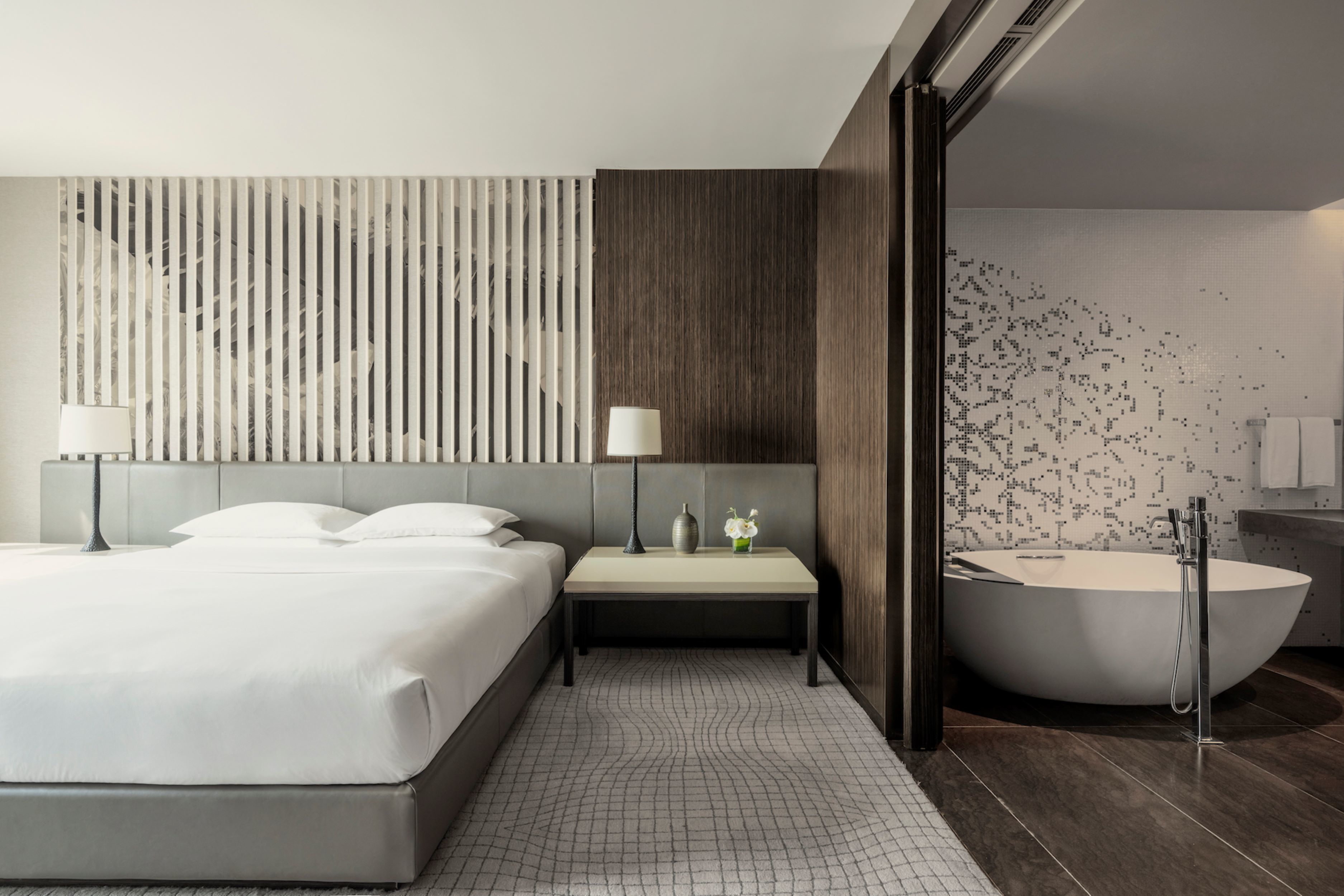 Una habitación de hotel moderna con una cama grande, una mesita de noche con lámpara, un jarrón pequeño con flores, una bañera amplia y una pared con textura.