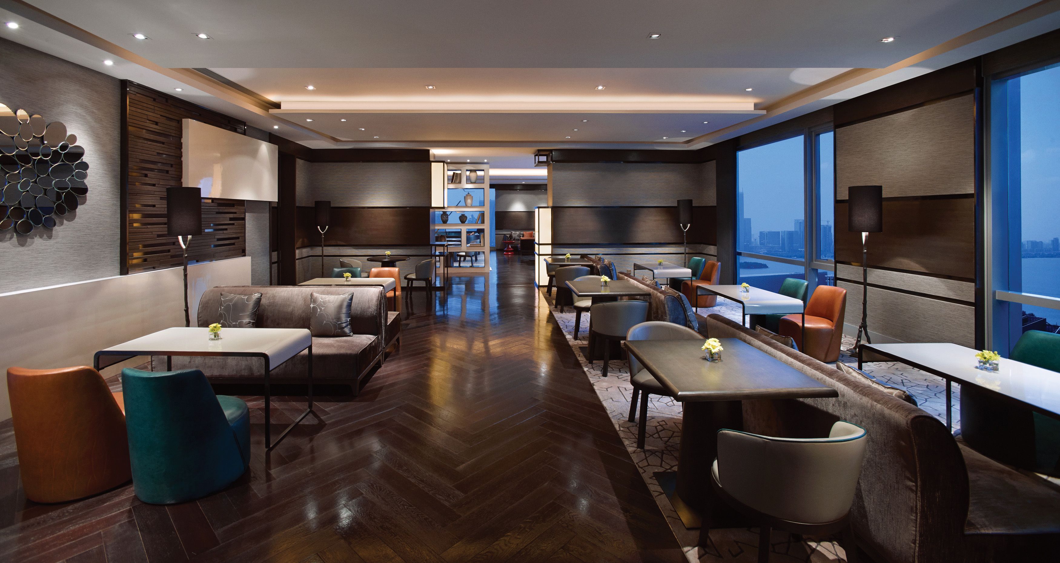 A modern hotel lobby with a herringbone wood floor, leather seating, and a city view through large windows.