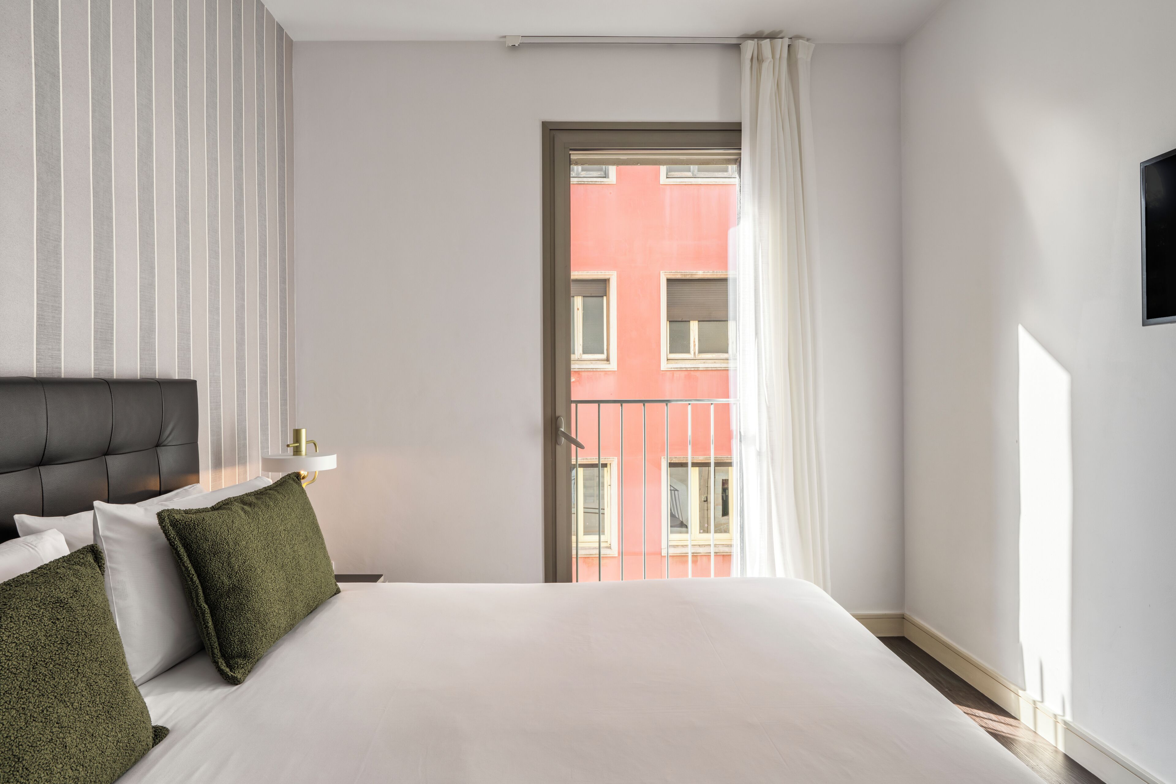Quarto de hotel com cama, mesa de cabeceira, abajur e vista para um edifício pela janela.