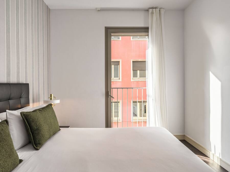 Quarto de hotel com cama, mesa de cabeceira, abajur e vista para um edifício pela janela.