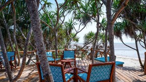 A wooden deck with chairs and tables, surrounded by palm trees, overlooking a beach.