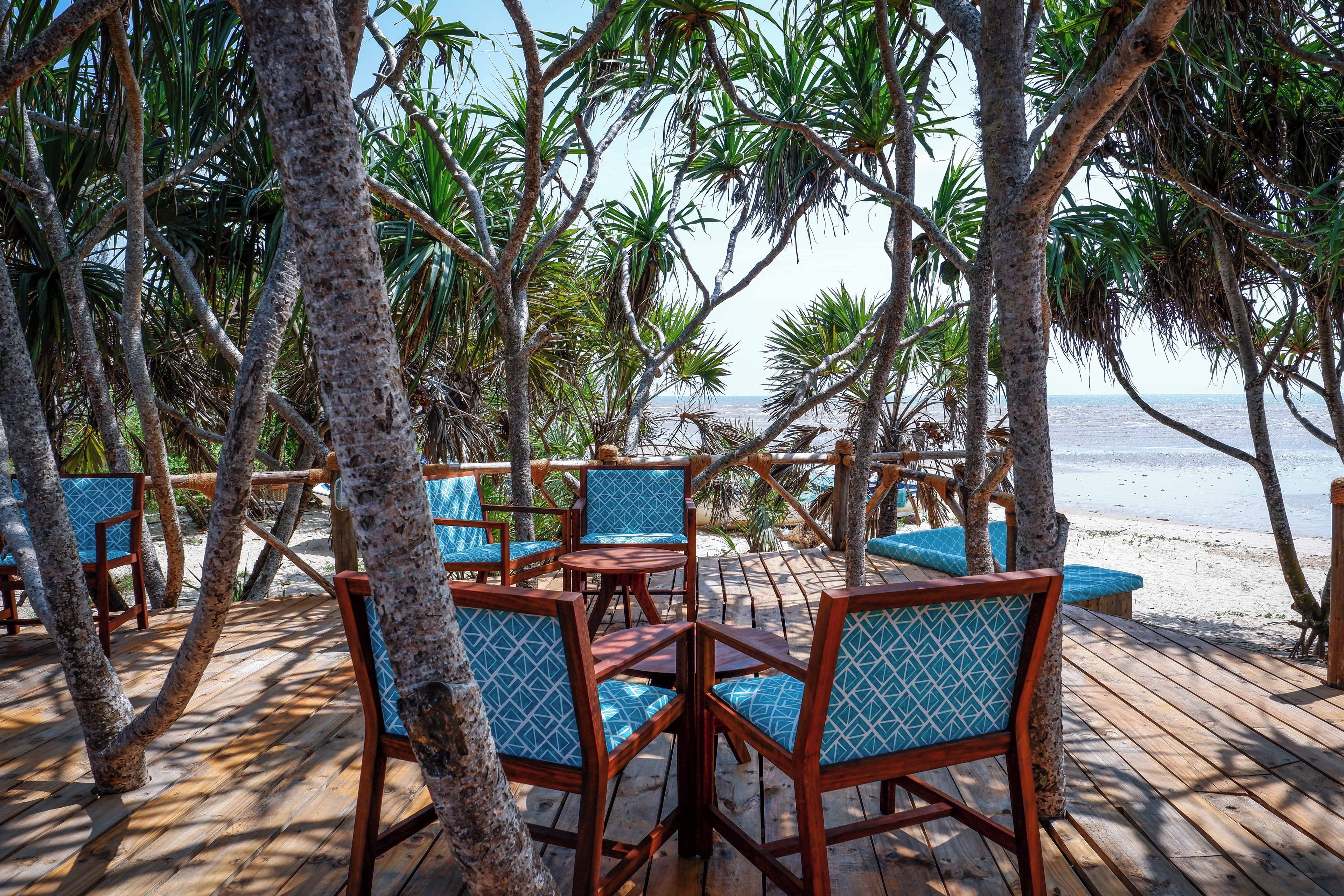 A wooden deck with chairs and tables, surrounded by palm trees, overlooking a beach.