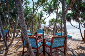 A wooden deck with chairs and tables, surrounded by palm trees, overlooking a beach. - Saadani Safari Lodge (Saadani)