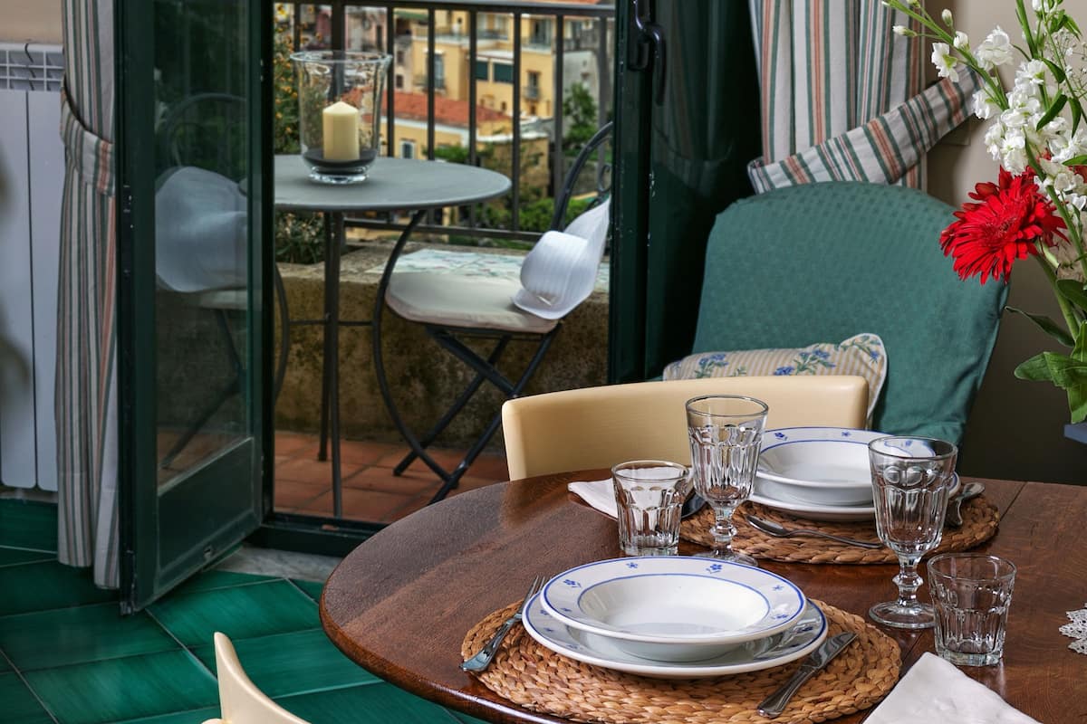Un coin repas avec une table dressée pour deux personnes, une vue sur le paysage urbain à travers une porte vitrée et une chaise verte.