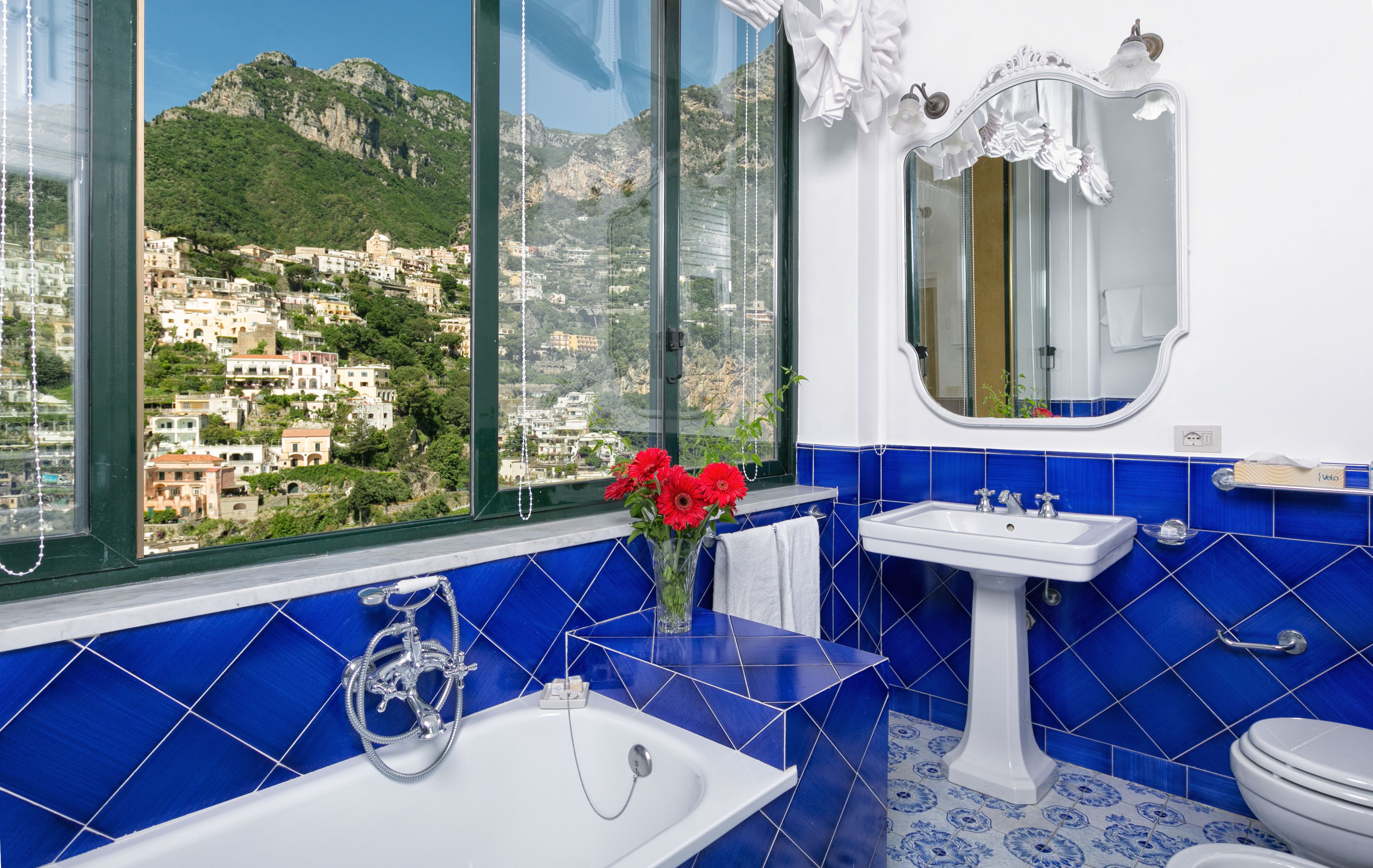 A bathroom with a bathtub, sink, and toilet, featuring blue tiles and a view of a mountainous town through large windows.
