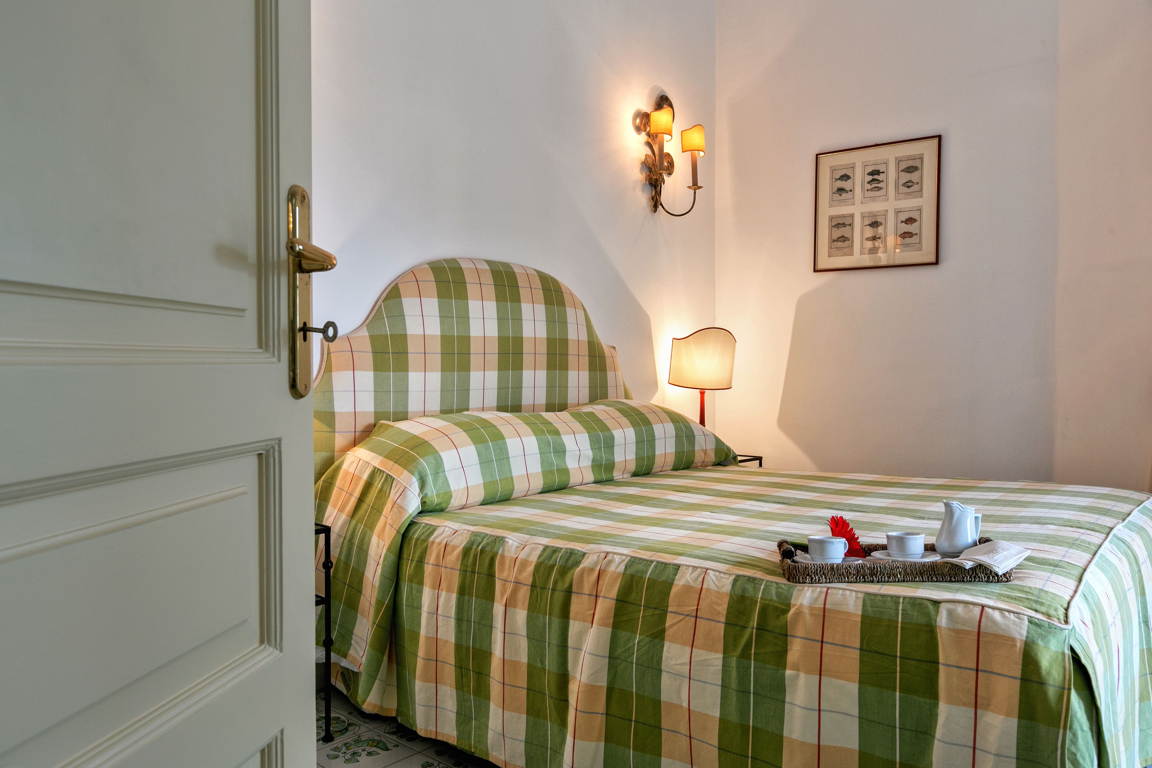 Een net opgemaakt bed met een geruit sprei, een bedlampje en een dienblad met een theepot, kopjes en een boek op het bed.