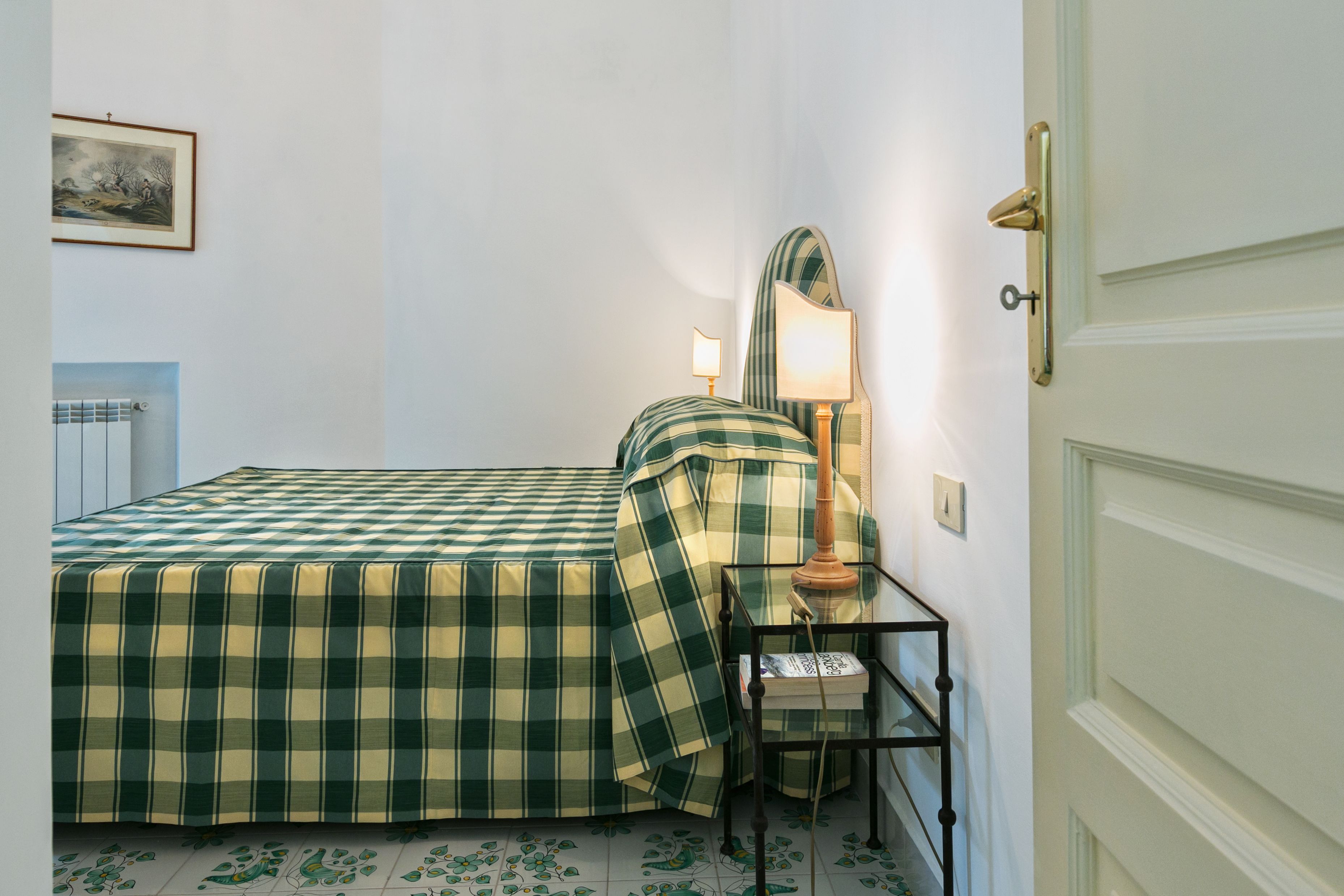 A room with a bed, a bedside table with a lamp, a framed picture on the wall, and a door.