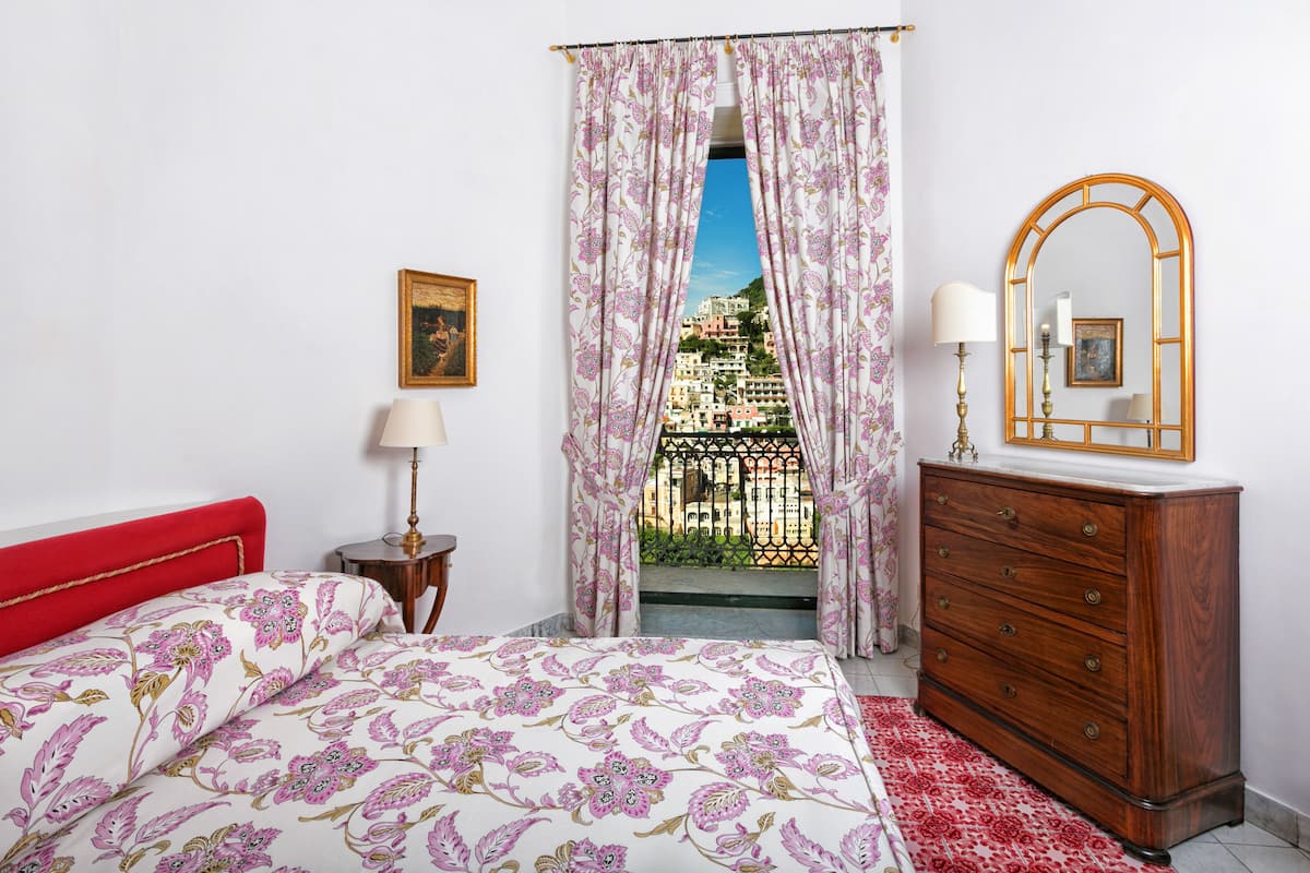 Une chambre à coucher avec un lit, une commode, un miroir et une vue sur les immeubles à travers les rideaux.