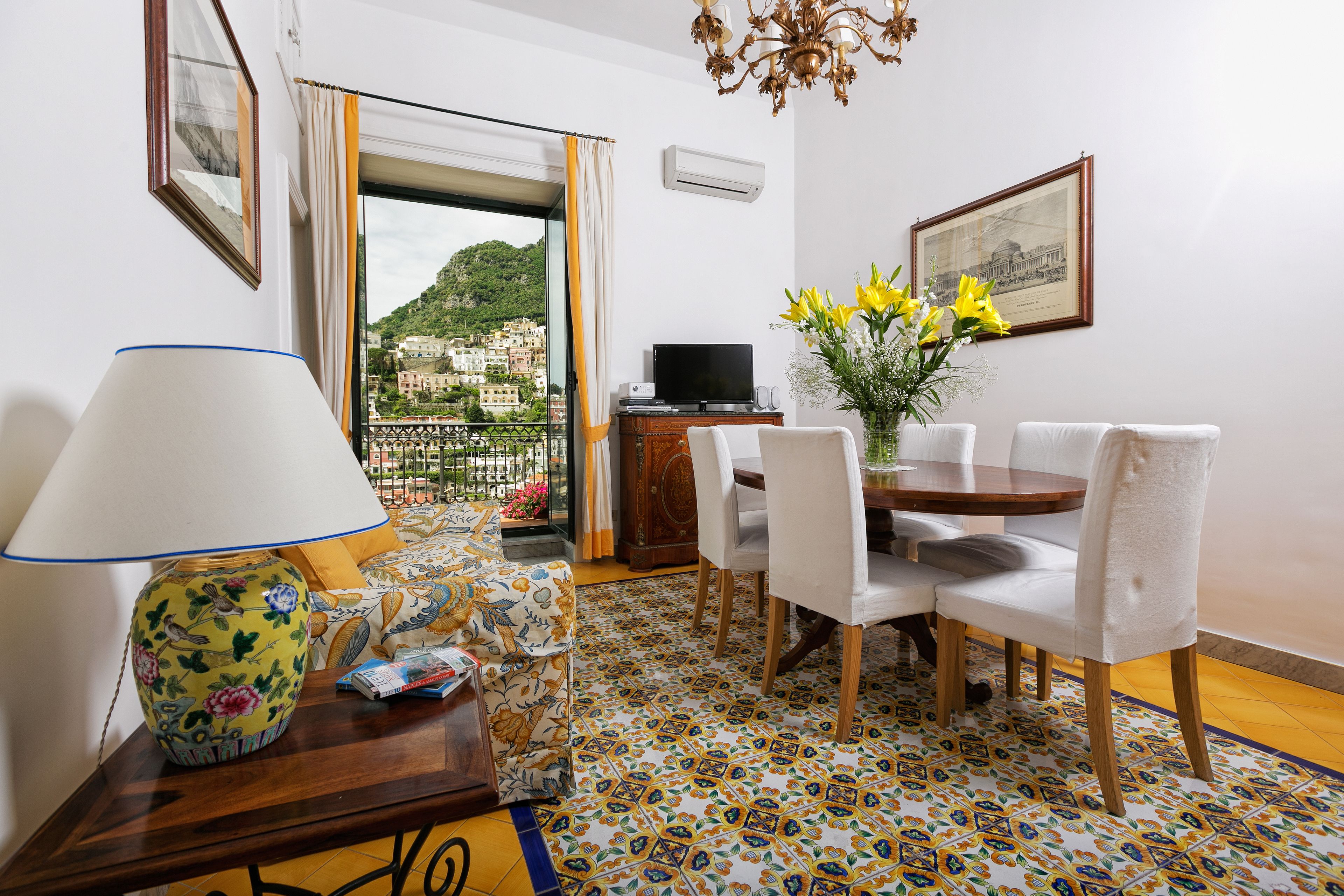 A dining area with a table, chairs, a TV, and a view of a hillside town.