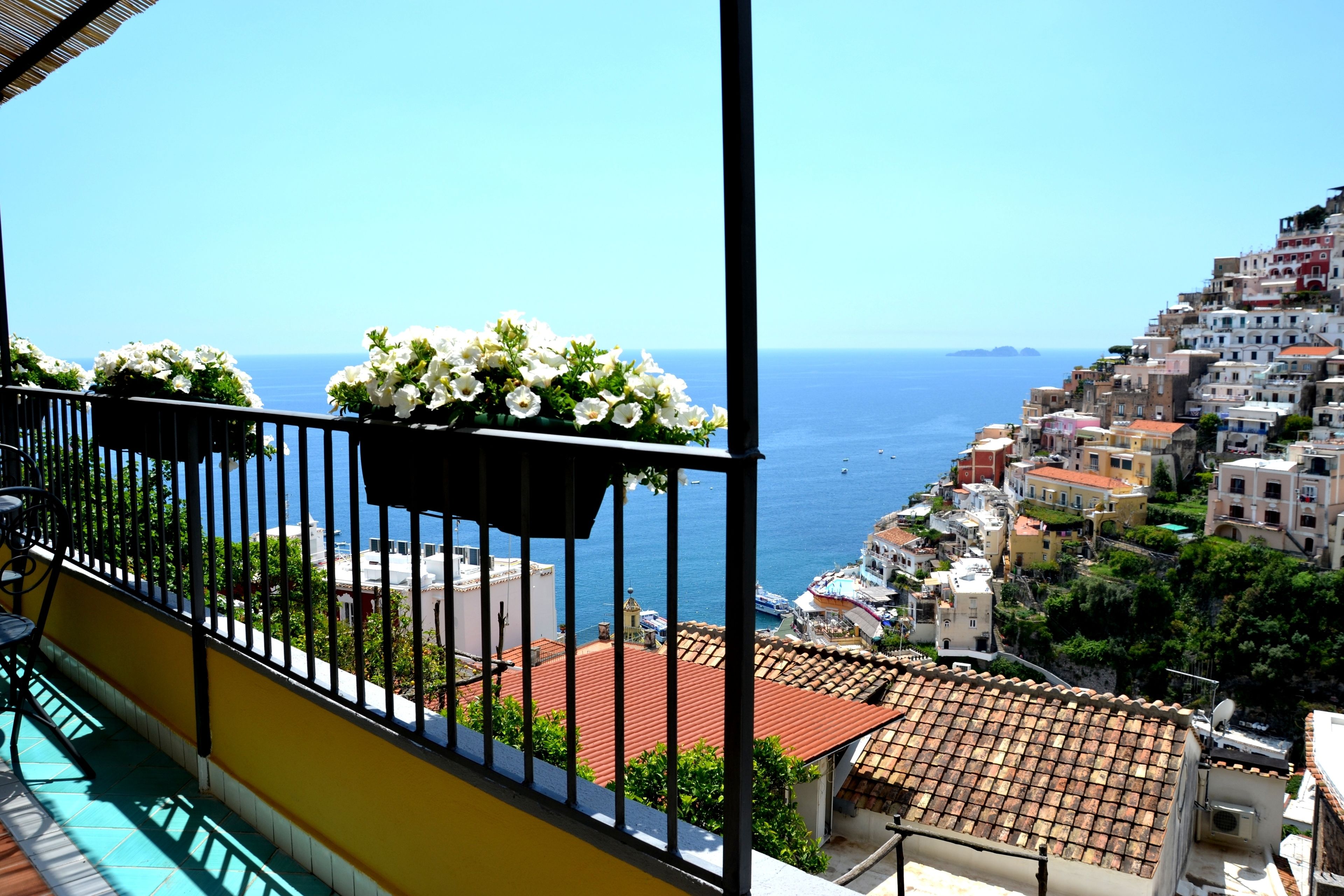 Un balcón con una maceta de flores, vista al mar y edificios con tejados de teja.