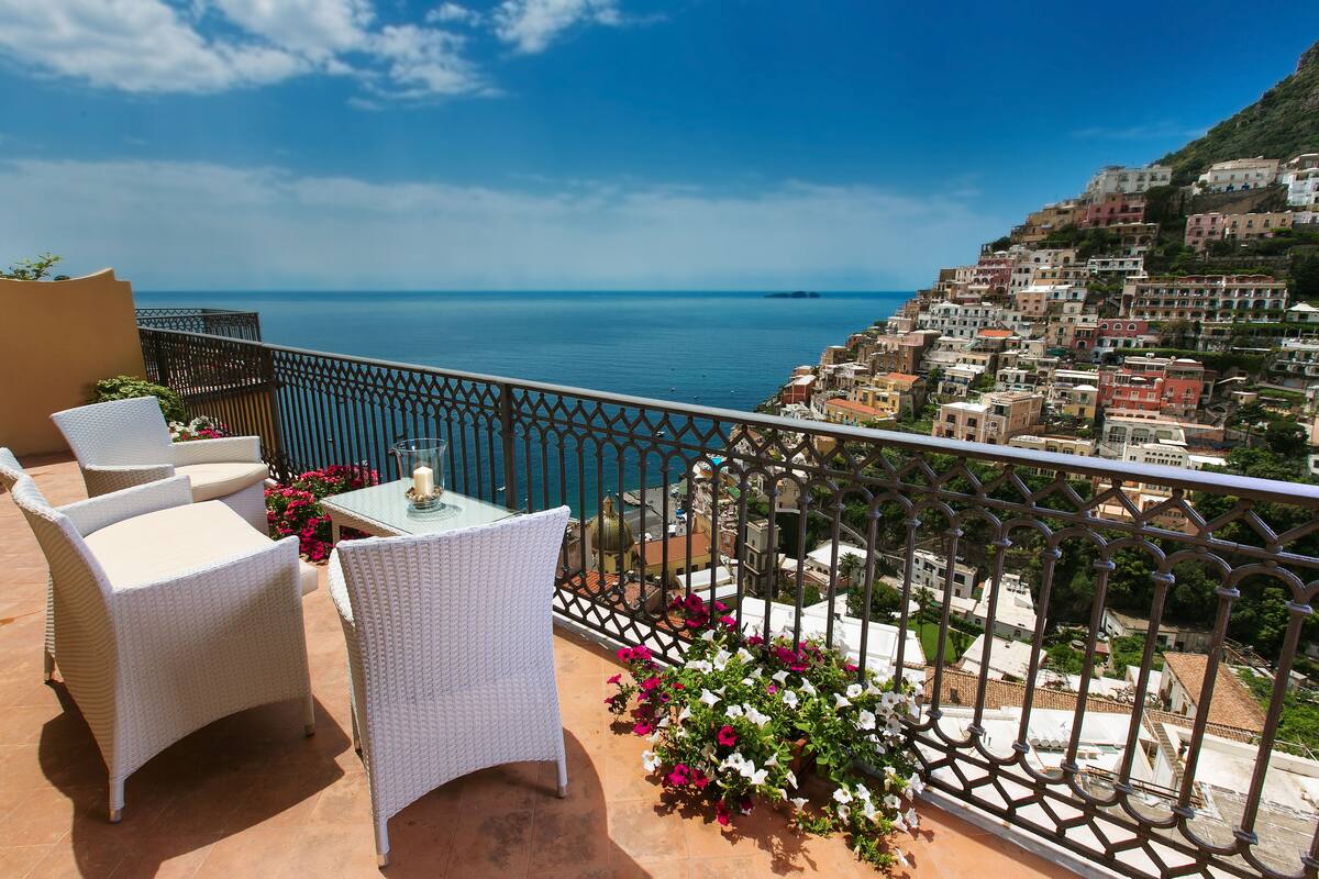 Une terrasse dotée de meubles en osier et d’une table, offrant une vue sur une ville côtière avec ses bâtiments et la mer.