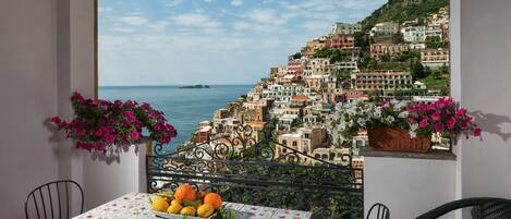 A balcony with a table and chairs, a flower arrangement, and a view of a coastal town.