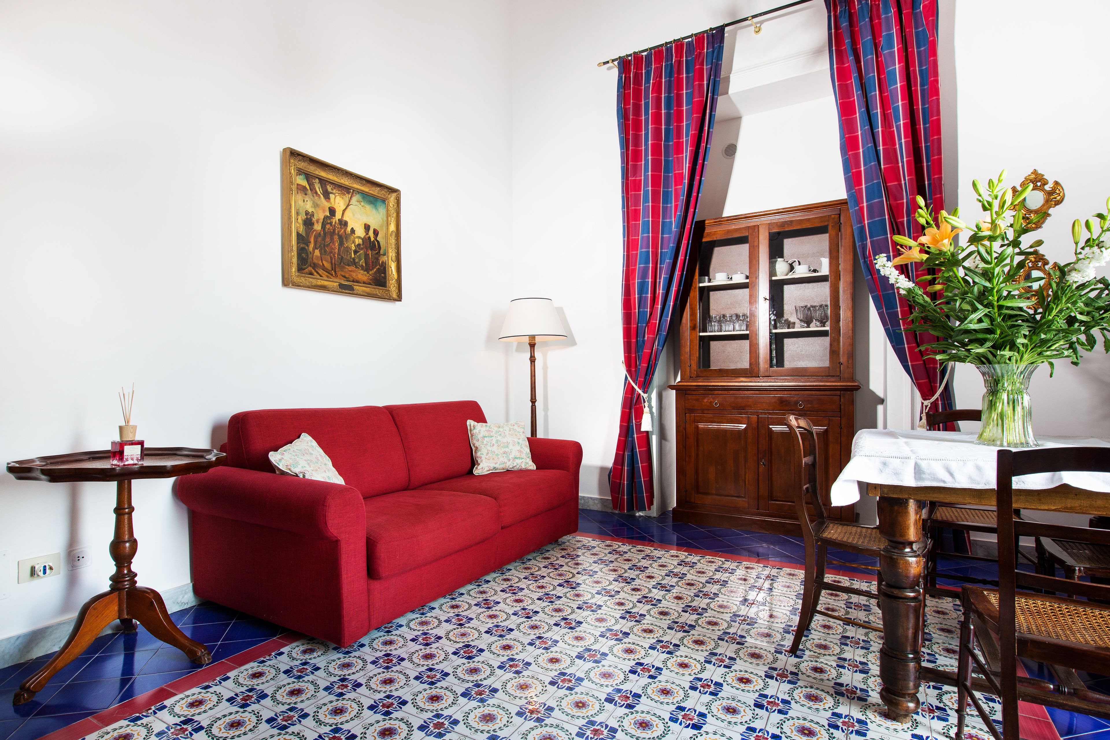 Un salon avec un canapé rouge, un meuble en bois, un tapis à motifs et une table sur laquelle est posé un vase de fleurs.