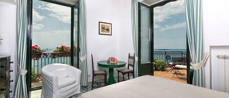 Habitación de hotel con cama, una mesita con sillas, balcón con vista al mar y una ventana con cortinas.