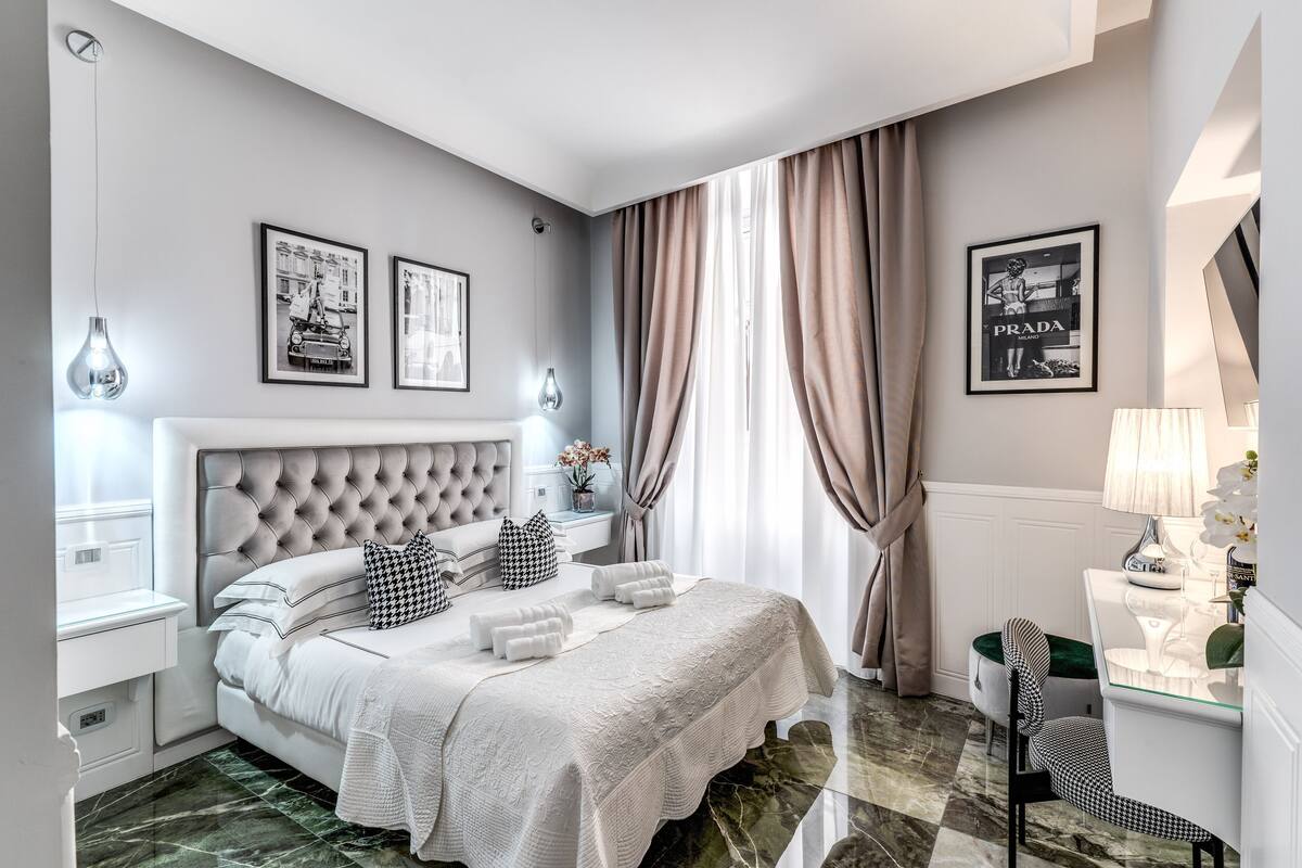 Un dormitorio moderno con un cabecero capitonado, una cama bien tendida con ropa de cama blanca, piso de mármol y una mesita de noche con lámpara.