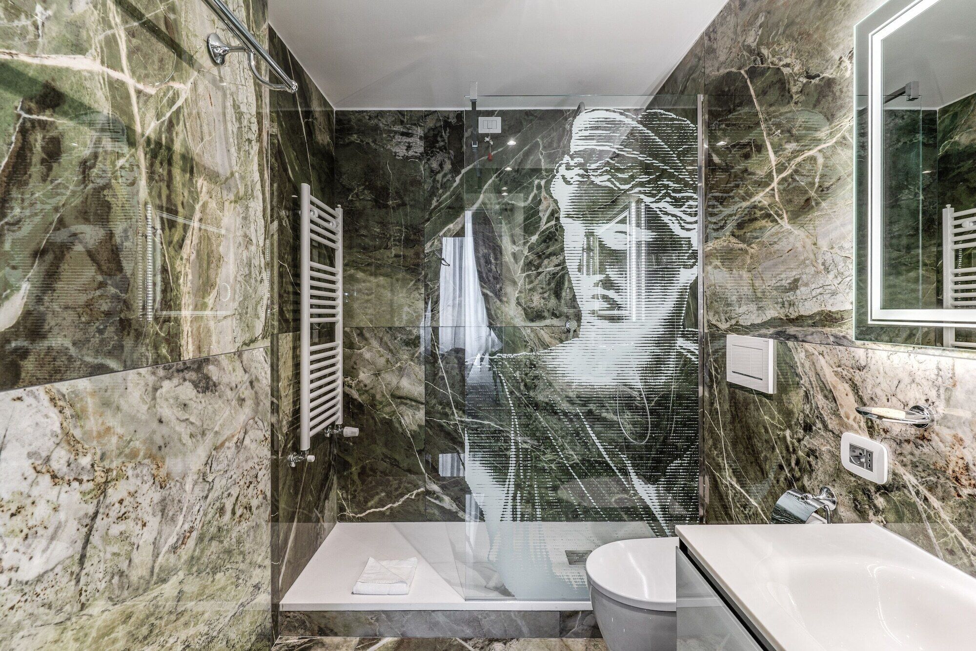 A modern bathroom with a glass shower enclosure, marble walls, and a large mirror.