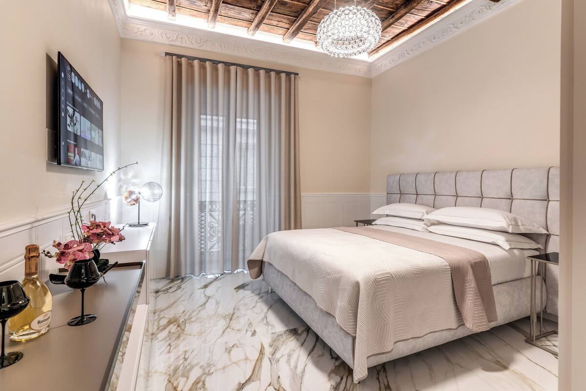 Un dormitorio moderno con una cama grande, suelo de mármol, televisión en la pared y una ventana con cortinas.