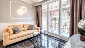 A modern living room with a beige sofa, a small side table, and a glass coffee table. There is a large window with curtains, a balcony visible outside, and a decorative wall piece. - Rome55 - Piazza di Spagna (Rome)