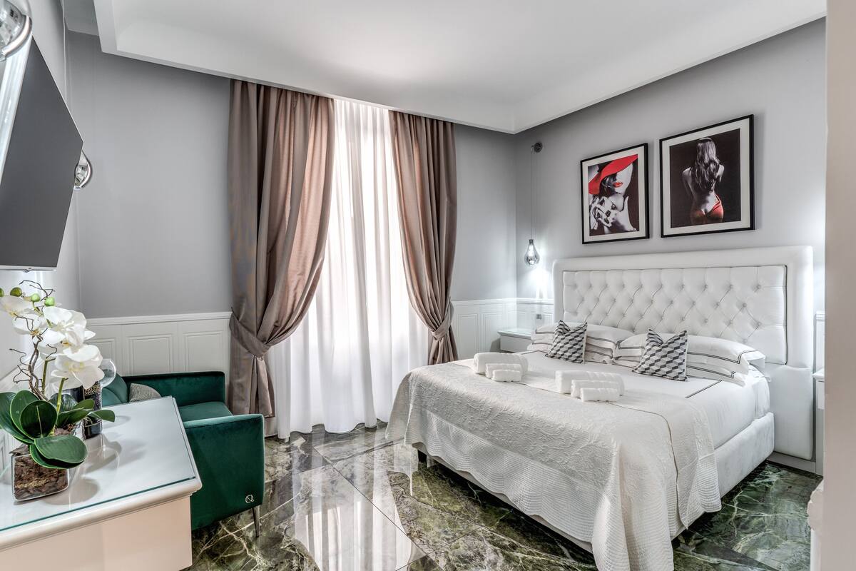 Una habitación de hotel moderna con una cama grande, un sillón verde, una mesa de centro de vidrio y una planta en maceta.