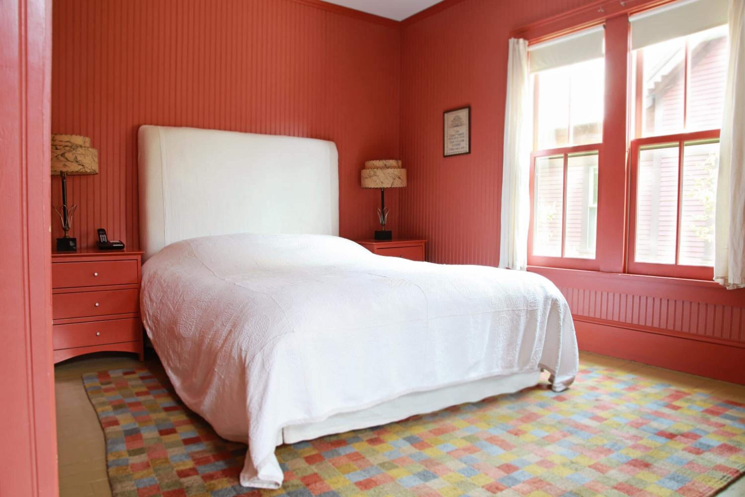 A bedroom with a large bed, two bedside tables, a window with shutters, and a colorful checkered floor.