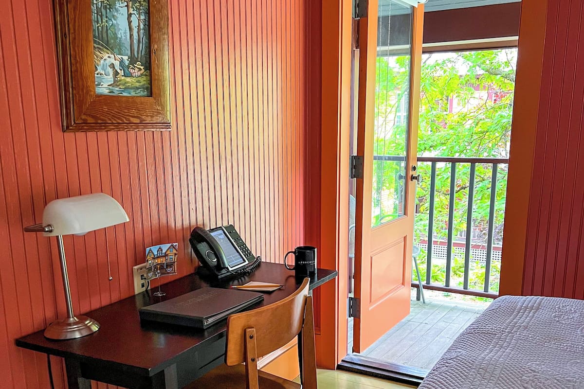 Une pièce comprenant un bureau en bois, une chaise, un lit, un tableau accroché au mur et une porte donnant sur un balcon extérieur.
