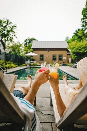 Two people relaxing by a poolside with drinks, a building in the background, and a clear sky.