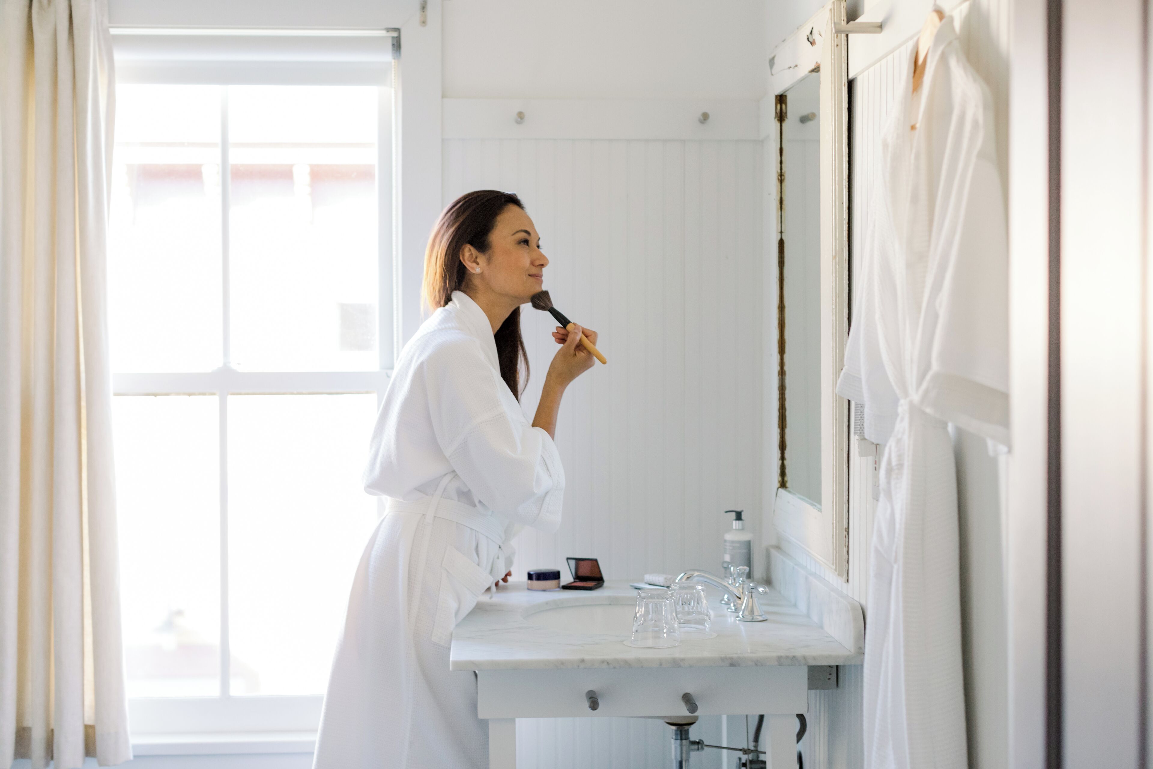 A person in a white robe standing in a bathroom with a mirror, a sink, and a window.