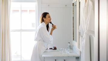 A person in a white robe standing in a bathroom with a mirror, a sink, and a window.