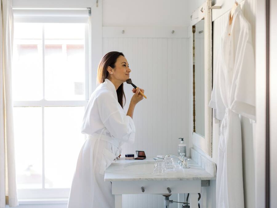 A person in a white robe standing in a bathroom with a mirror, a sink, and a window.
