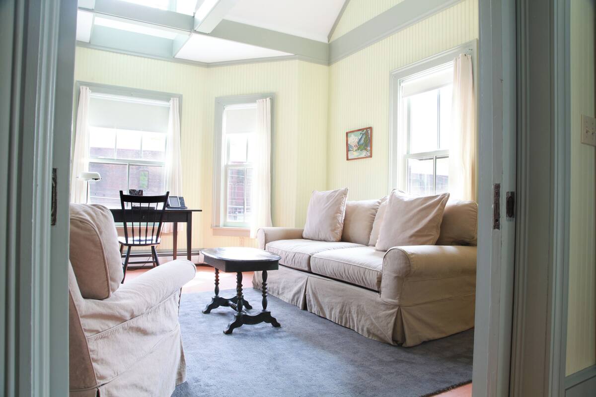 Un salon avec un canapé beige, une petite table ronde et un fauteuil. Il y a deux fenêtres avec des rideaux, et un tableau accroché au mur.