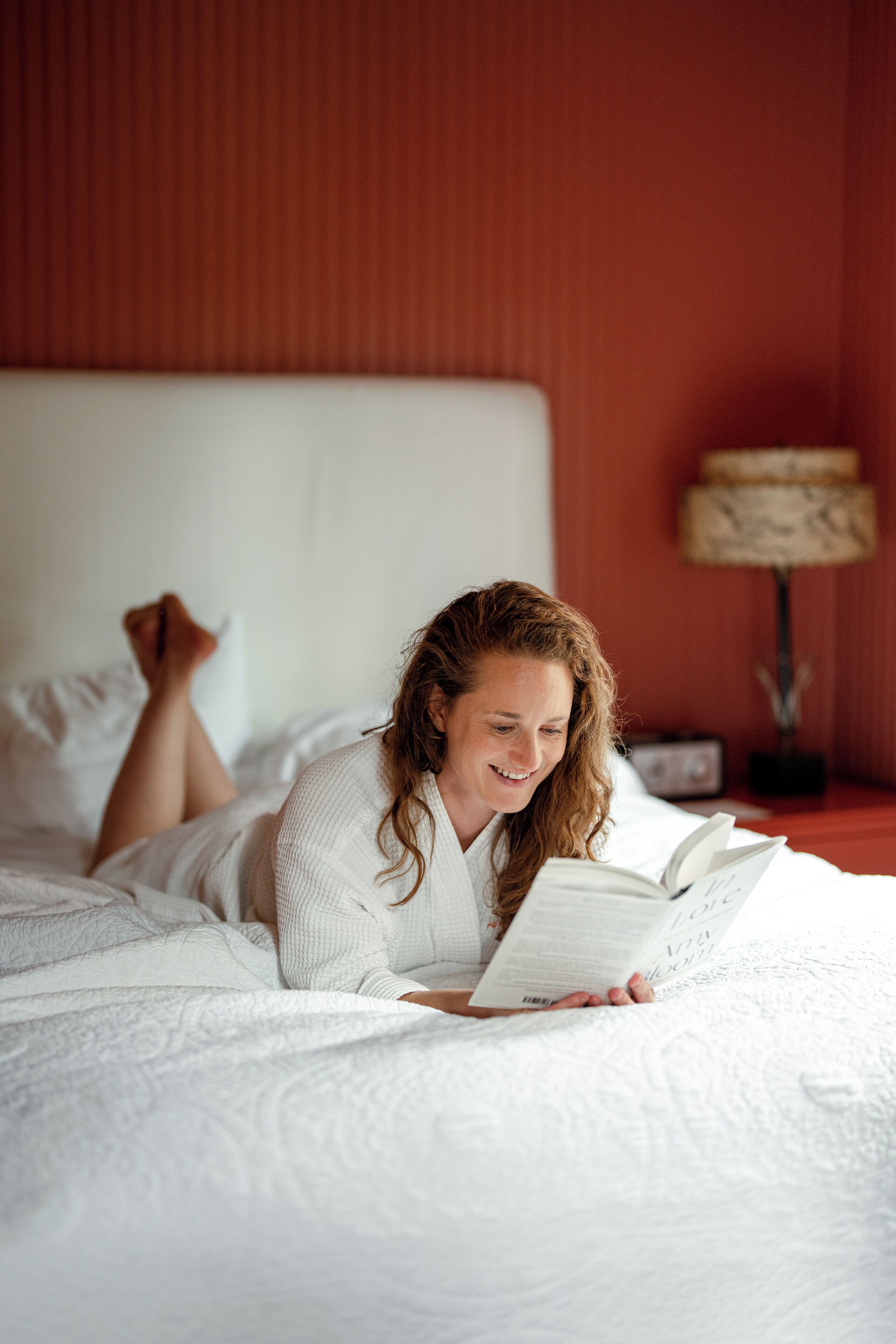 A person lying on a bed with a book, in a room with a red wall and a lamp in the background.