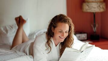A person lying on a bed with a book, in a room with a red wall and a lamp in the background.