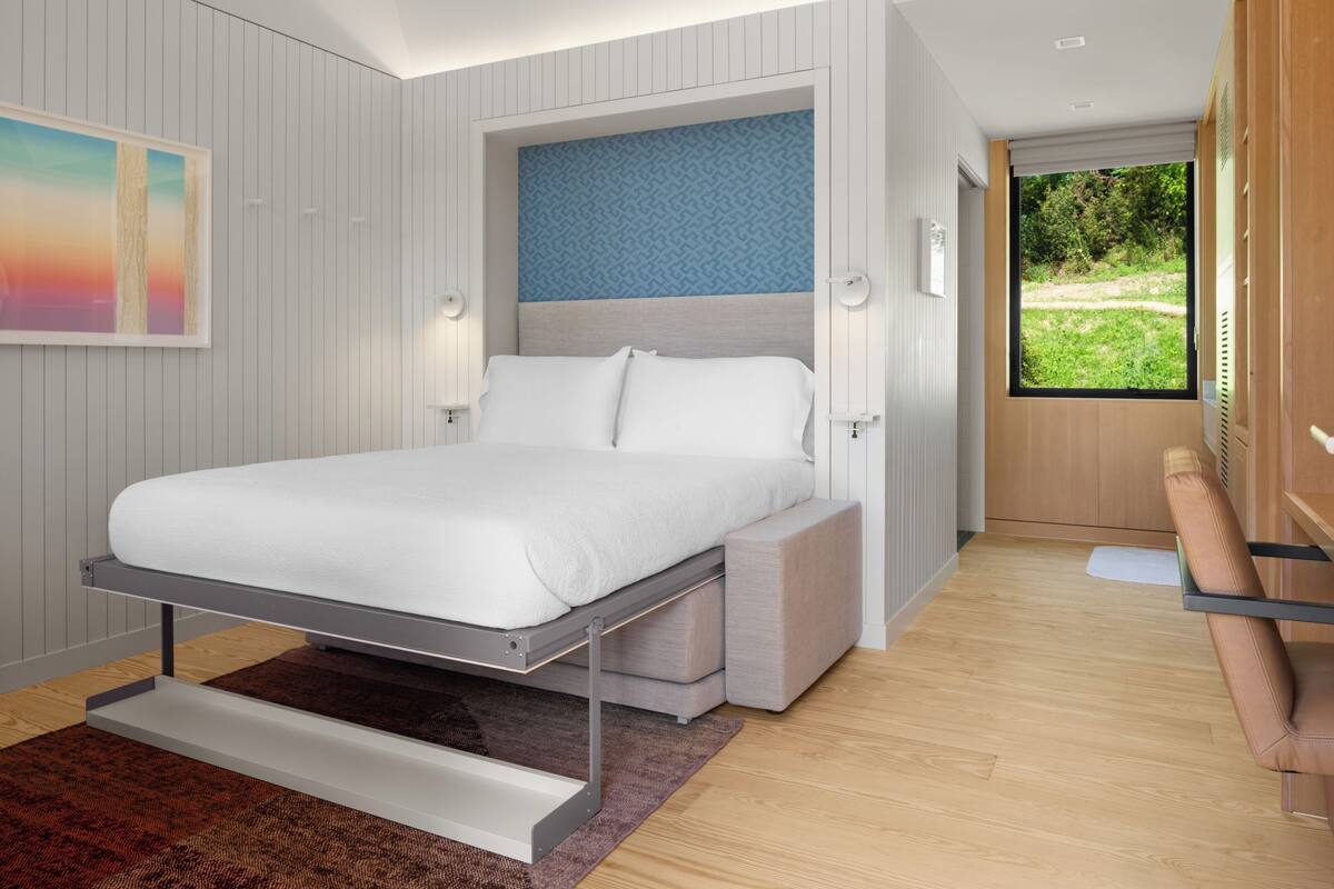 Une chambre moderne avec un lit, un banc intégré et une grande fenêtre donnant sur un paysage verdoyant.