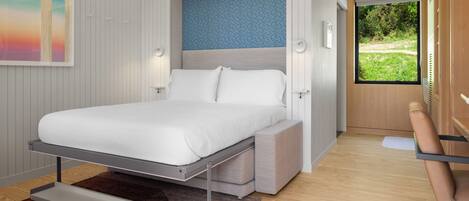 Un dormitorio moderno con cama, banco empotrado y un amplio ventanal con vistas a la vegetación.