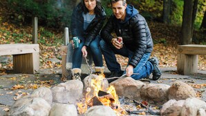 Two people sitting by a campfire in a wooded area with benches and a clear view of the surrounding trees. - The Porches Inn at Mass MoCA (North Adams)